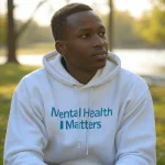 Person wearing a white hoodie with "Mental Health Matters" text, seated in a park with trees and water in the background.